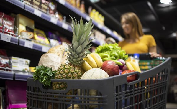 Close up view of shopping cart overloaded with food while in background female person choosing products.
