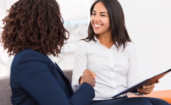 Happy diverse business partners closing deal. Businesswomen sitting on couch, holding folder with documents, shaking hands and smiling. Agreement concept